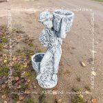 Светло-серая бетонная скульптура мальчика с двумя корзинками, украшенными рельефным узором; фигура стоит на дорожке.