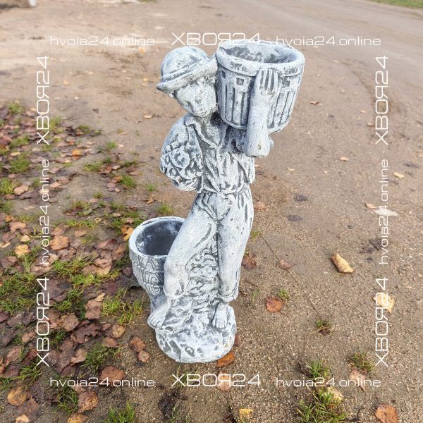 Светло-серая бетонная скульптура мальчика с двумя корзинками, украшенными рельефным узором; фигура стоит на дорожке.