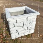 Белая квадратная цветочница из бетона с широким бортиком и рельефной фактурой кирпичной кладки стоит на земле.