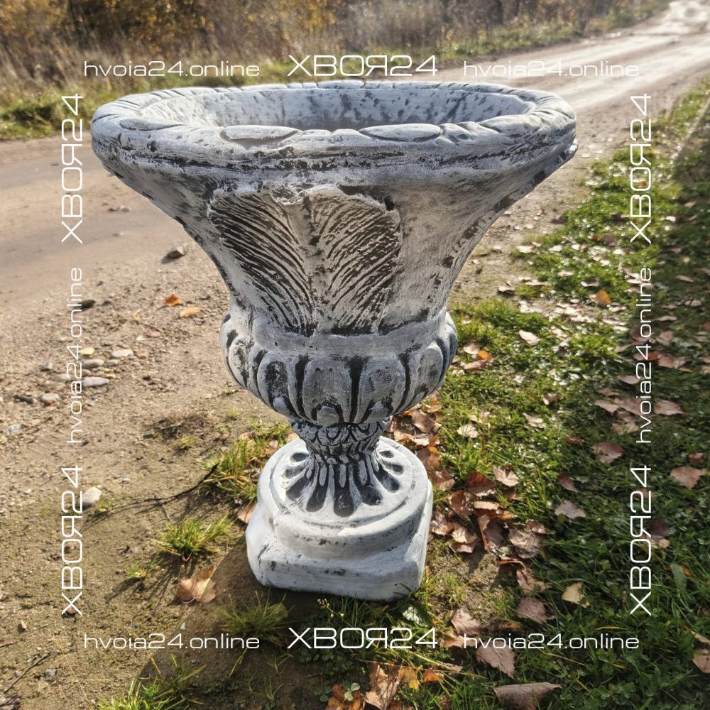Бетонный вазон М035, рельефная чаша на фигурной ножке Серый бетонный вазон М035 с широкой чашей и рельефными узорами на фигурной ножке стоит у дорожки среди травы и опавшей листвы.