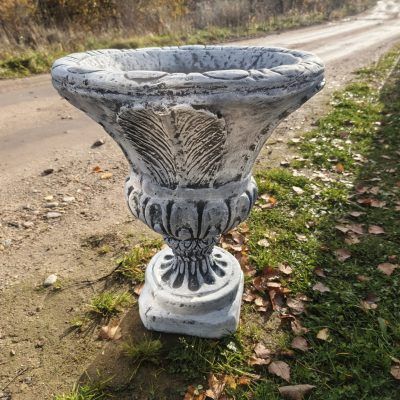 Серый бетонный вазон М035 с широкой чашей и рельефными узорами на фигурной ножке стоит у дорожки среди травы и опавшей листвы.
