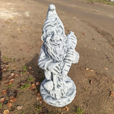 Светло-серый бетонный садовый гном с длинной бородой держит крупную морковь; фигурка стоит на земле среди опавших листьев.
